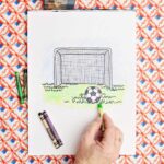 A person putting finishing coloring touches on a drawing of a soccer goal and ball in the grass.