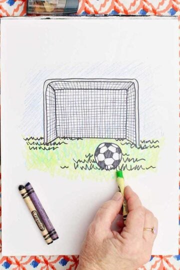 A person putting finishing coloring touches on a drawing of a soccer goal and ball in the grass.