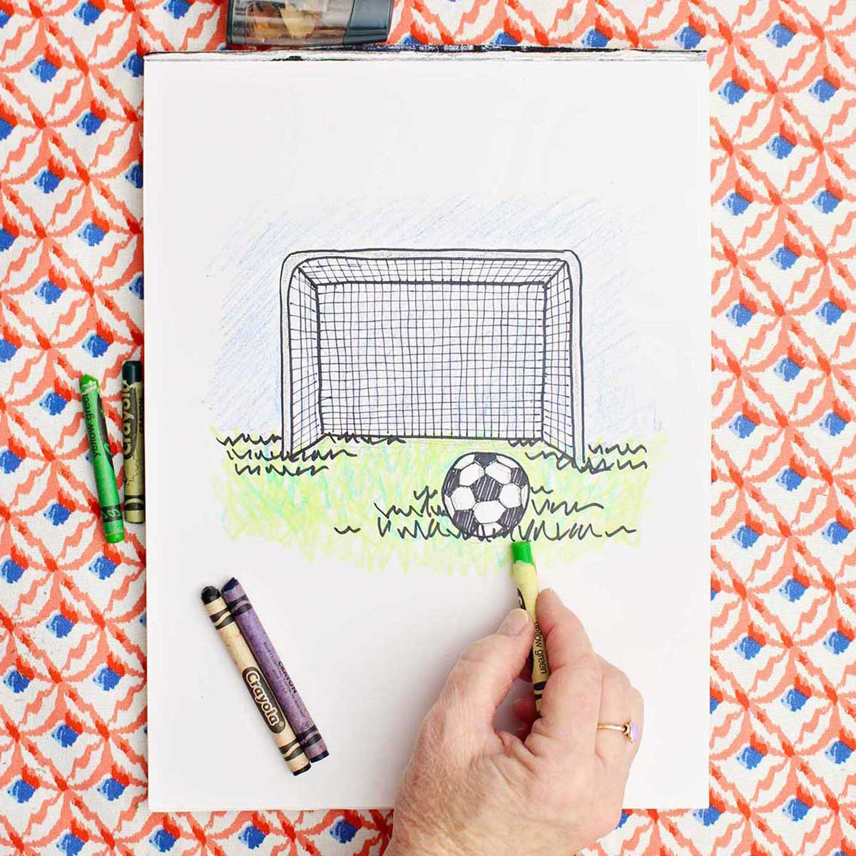 A person putting finishing coloring touches on a drawing of a soccer goal and ball in the grass.