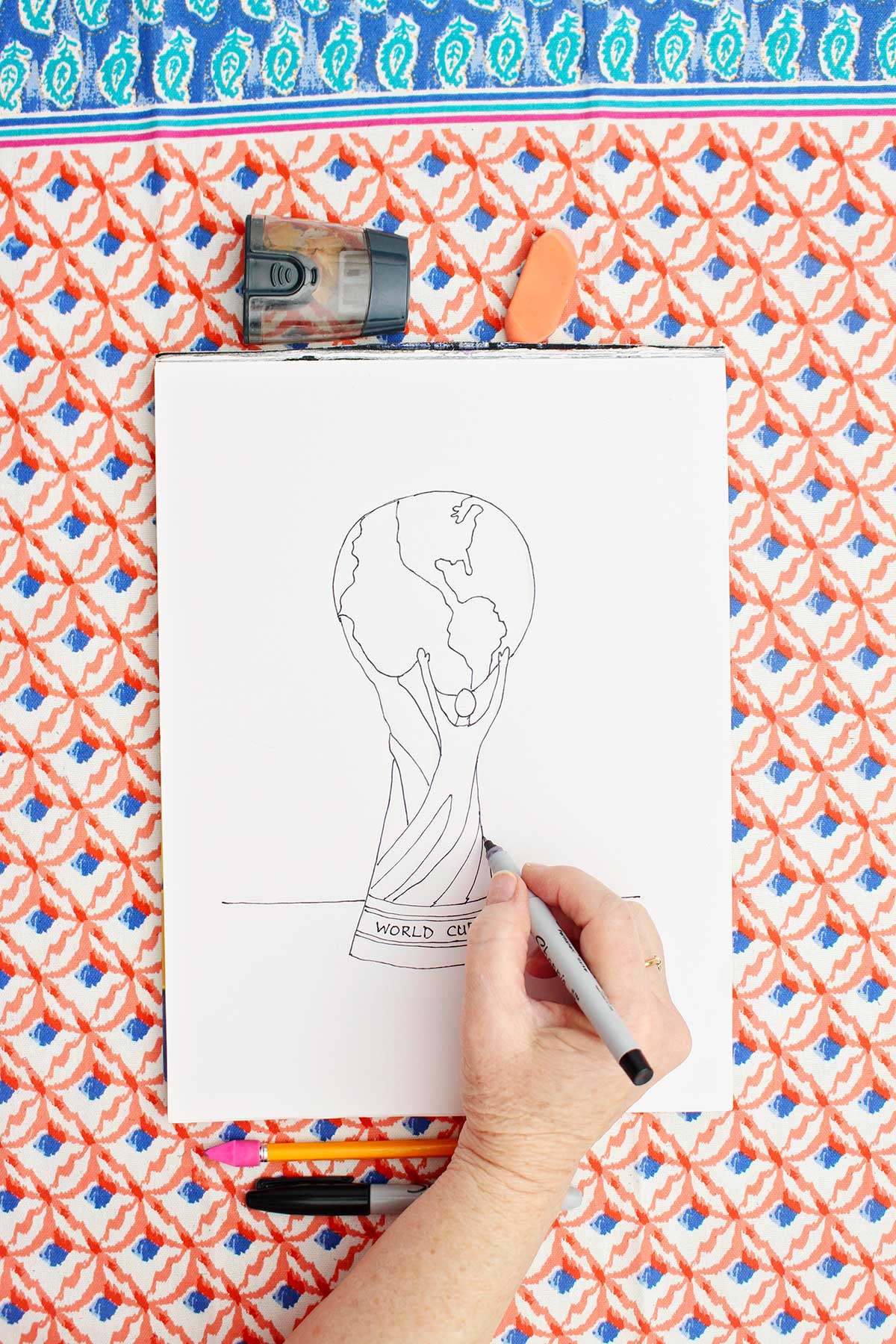 A person going over a sketched drawing of the FIFA World Cup trophy with sharpie marker.