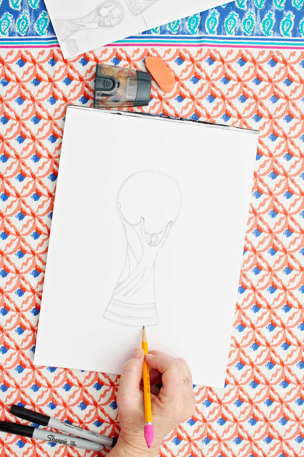 A person adding a base to the bottom of a pencil sketch of the FIFA World Cup trophy.