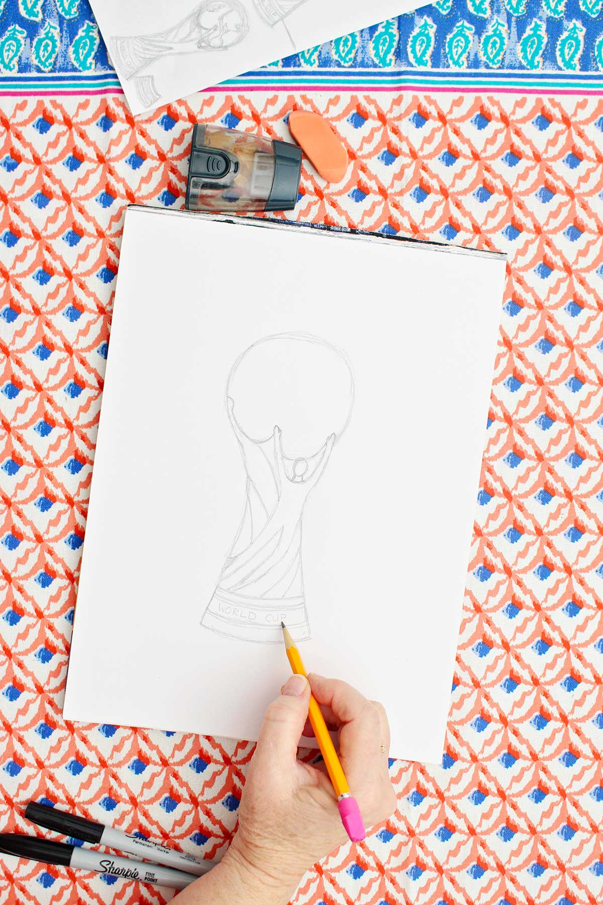 A person adding text to the bottom of a pencil sketch of the FIFA World Cup trophy.