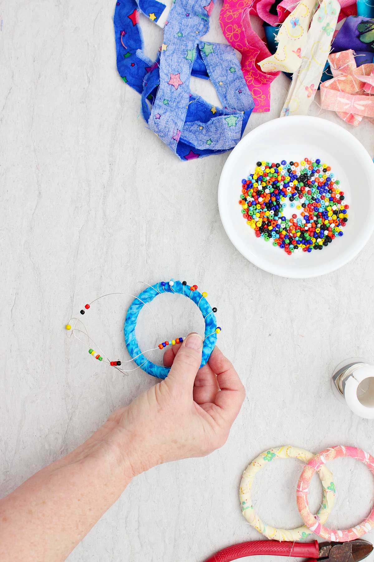 A person wrapping a bead and wire on a fabric BoHo bracelet with other supplies near by.