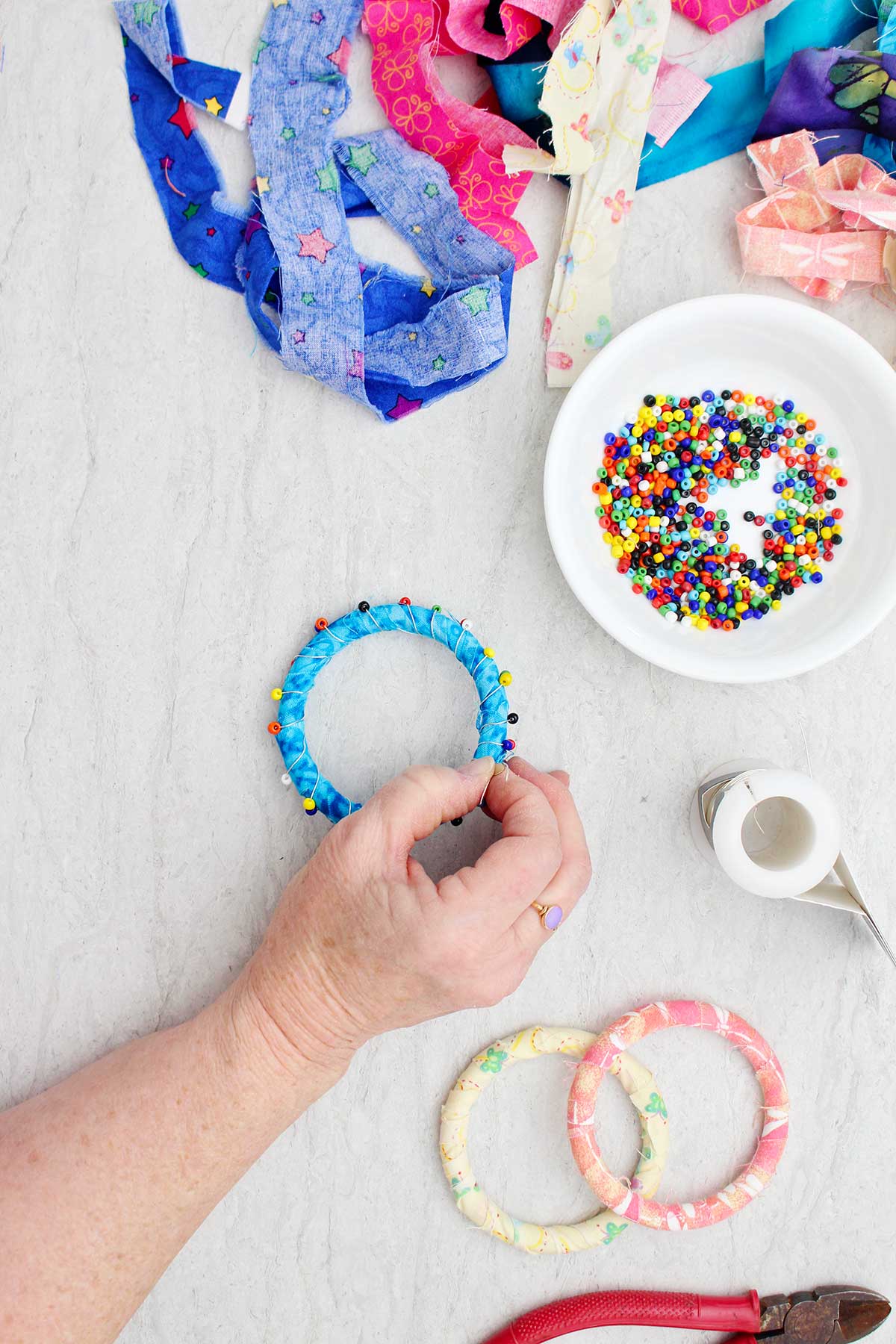 A person securing a bead and wire wrap on a fabric BoHo bracelet with other supplies near by.