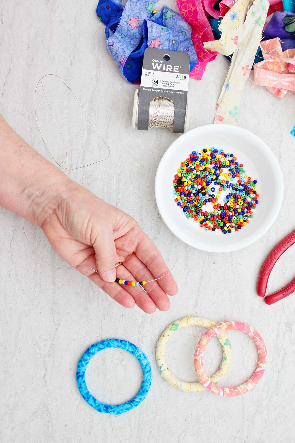 A person slipping colorful small beads on a wire with BoHo bracelets and strips of fabric near by.