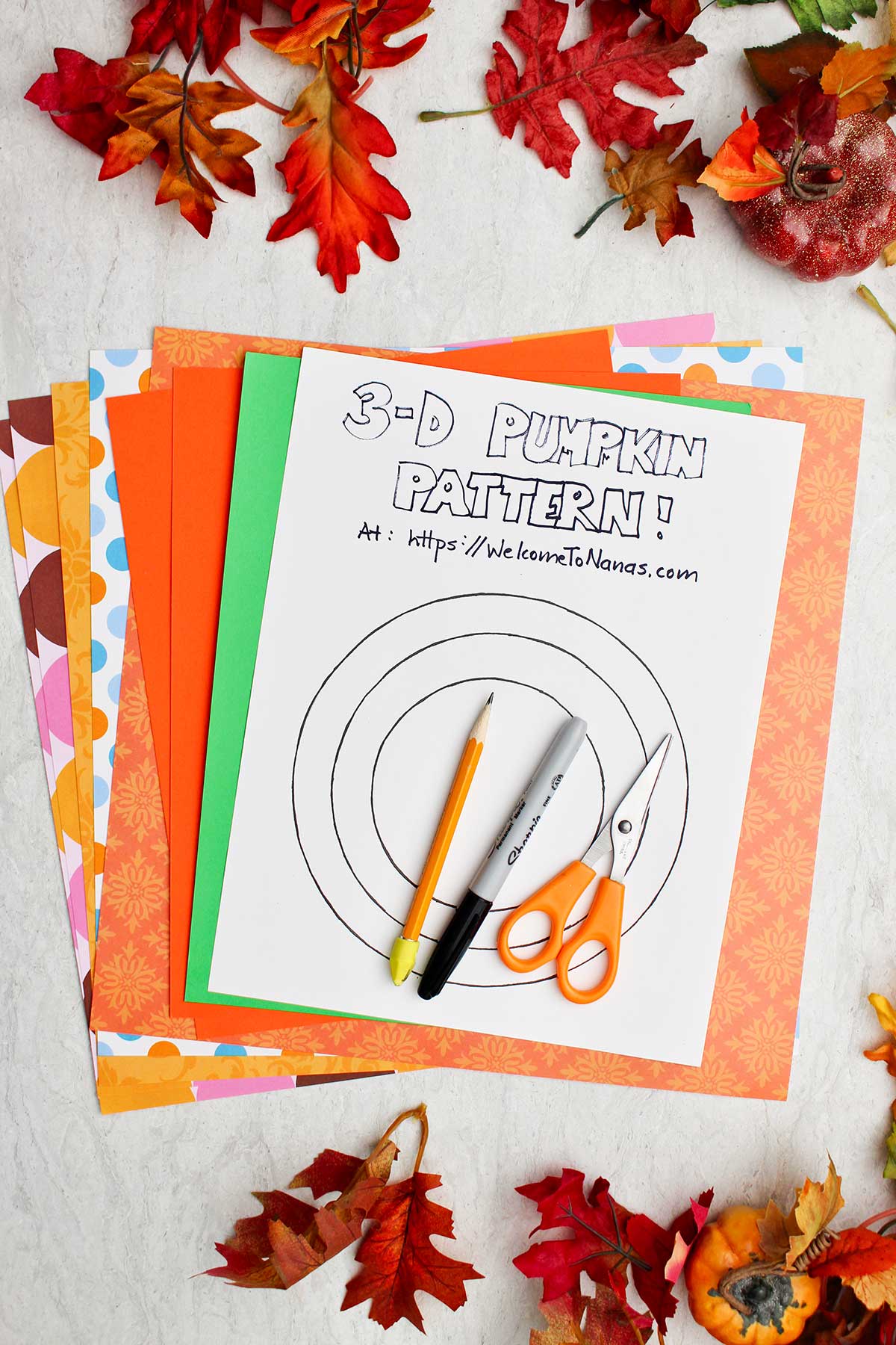 A pile of colorful patterned paper, a 3D pumpkin pattern, a pencil a sharpie and scissors.