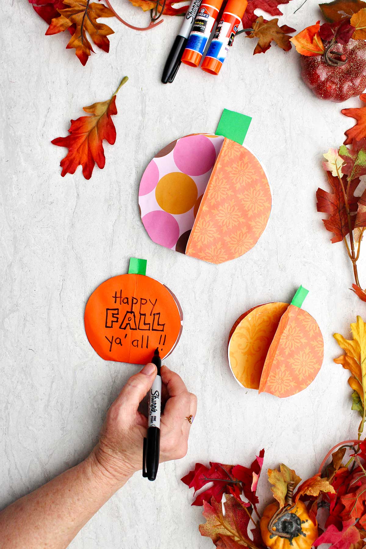 A person writing a note "Happy Fall Ya'all" on a pumpkin card with two other 3D pumpkins and fall leaves near by.