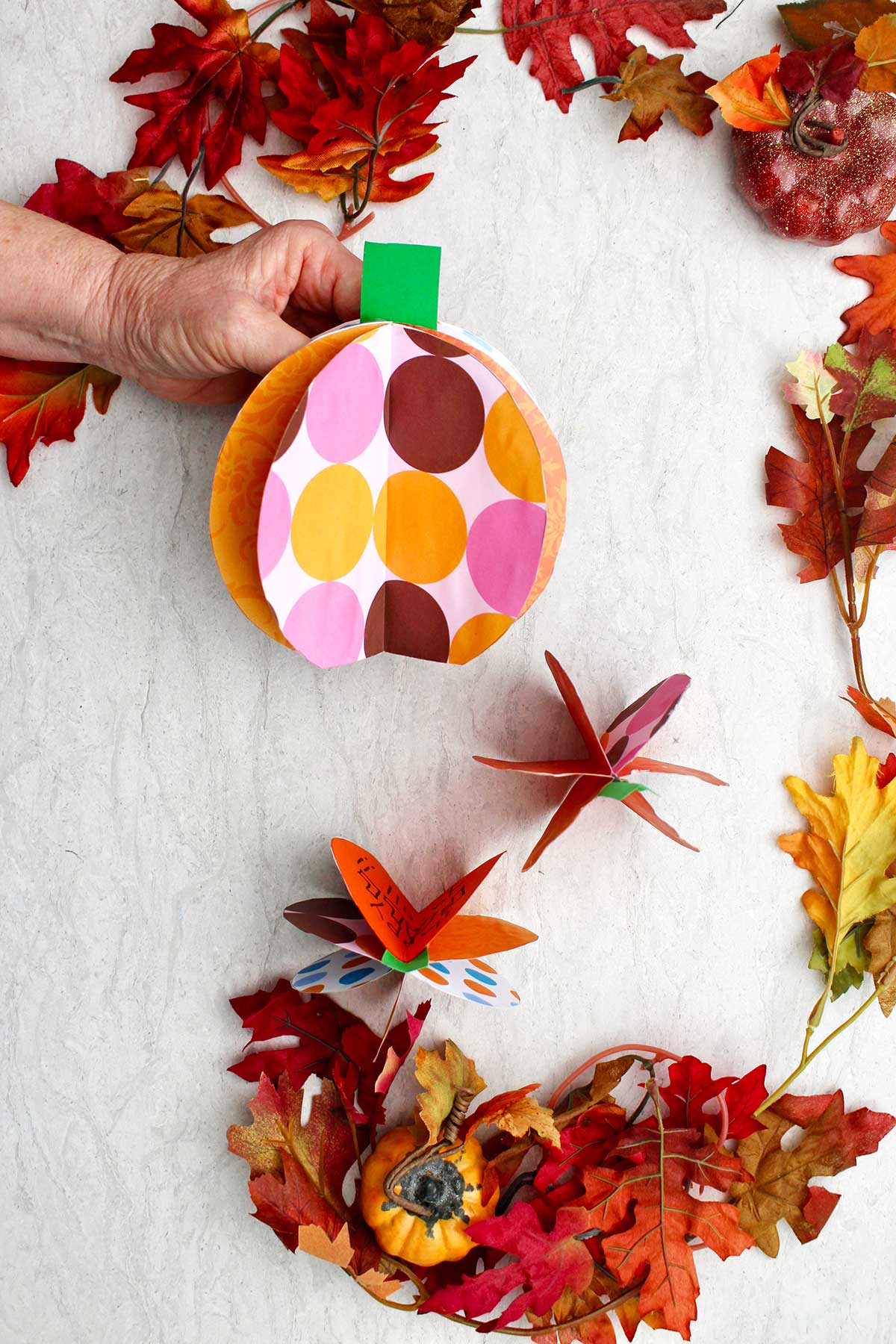 A person holding a 3D paper pumpkin with fall leaves scattered around.