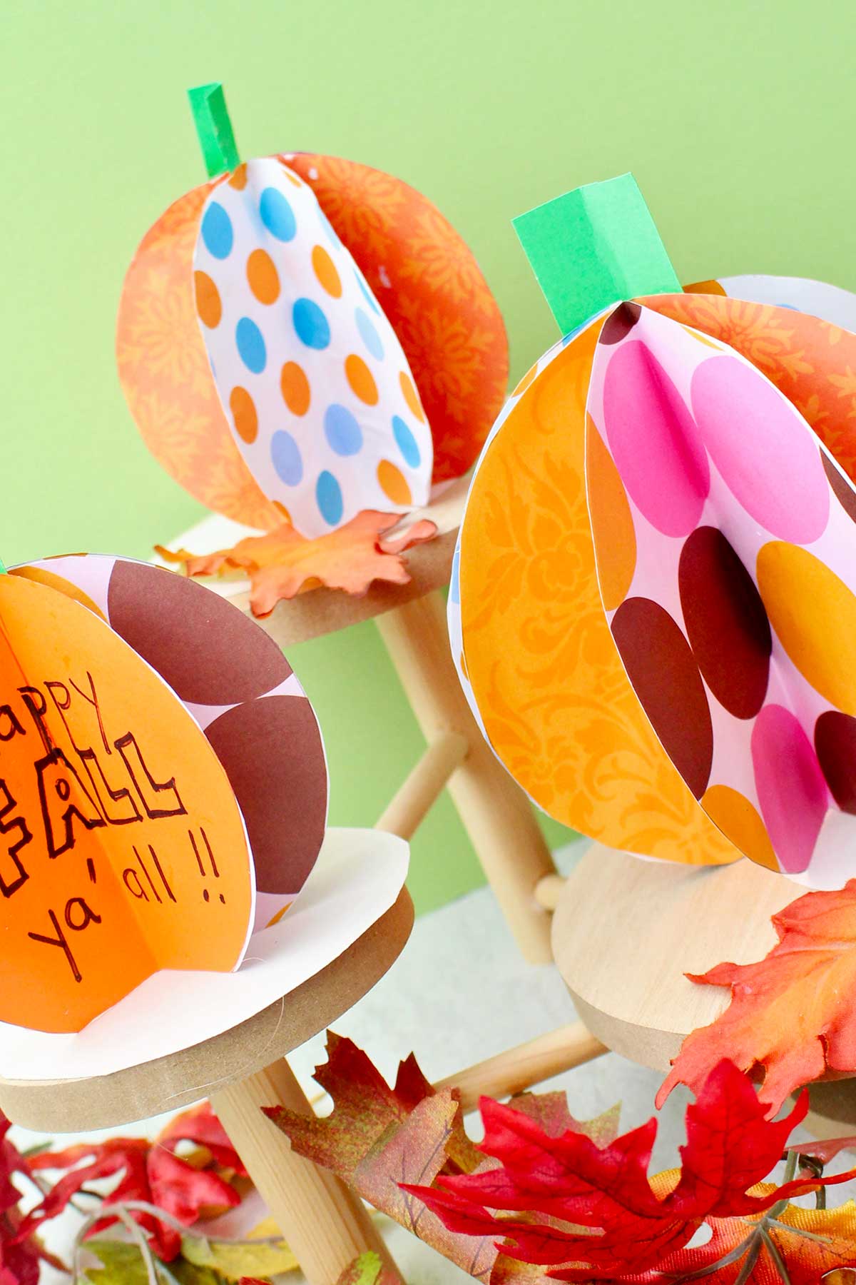 Three colorful paper pumpkins on wooden pedestals with fake leaves decorating near by.