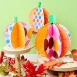 Three colorful paper pumpkins on wooden pedestals with fake leaves decorating near by.