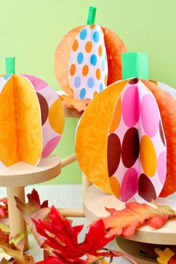 Three colorful paper pumpkins on wooden pedestals with fake leaves decorating near by.