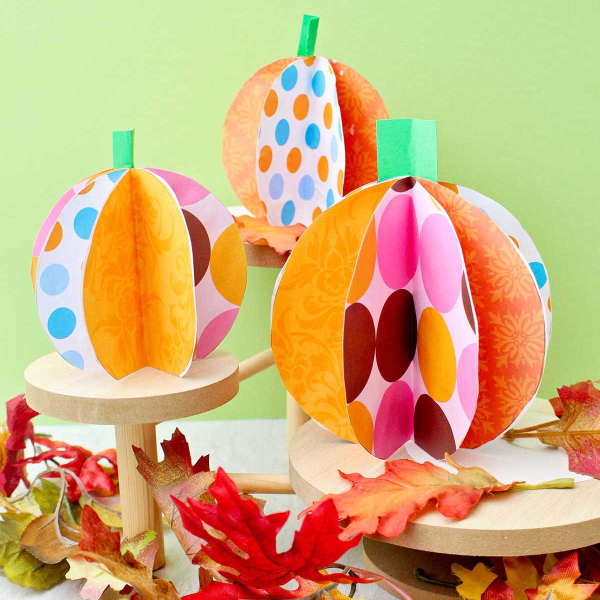 Three colorful paper pumpkins on wooden pedestals with fake leaves decorating near by.