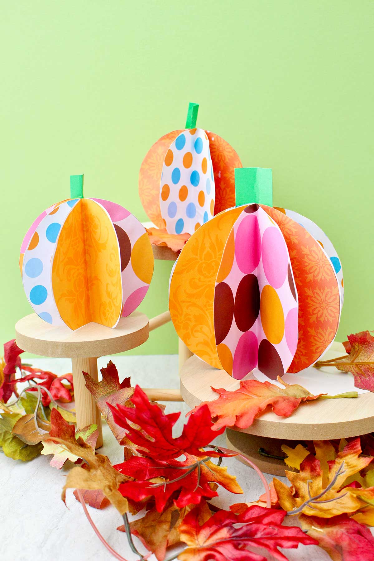 Three colorful paper pumpkins on wooden pedestals with fake leaves decorating near by.