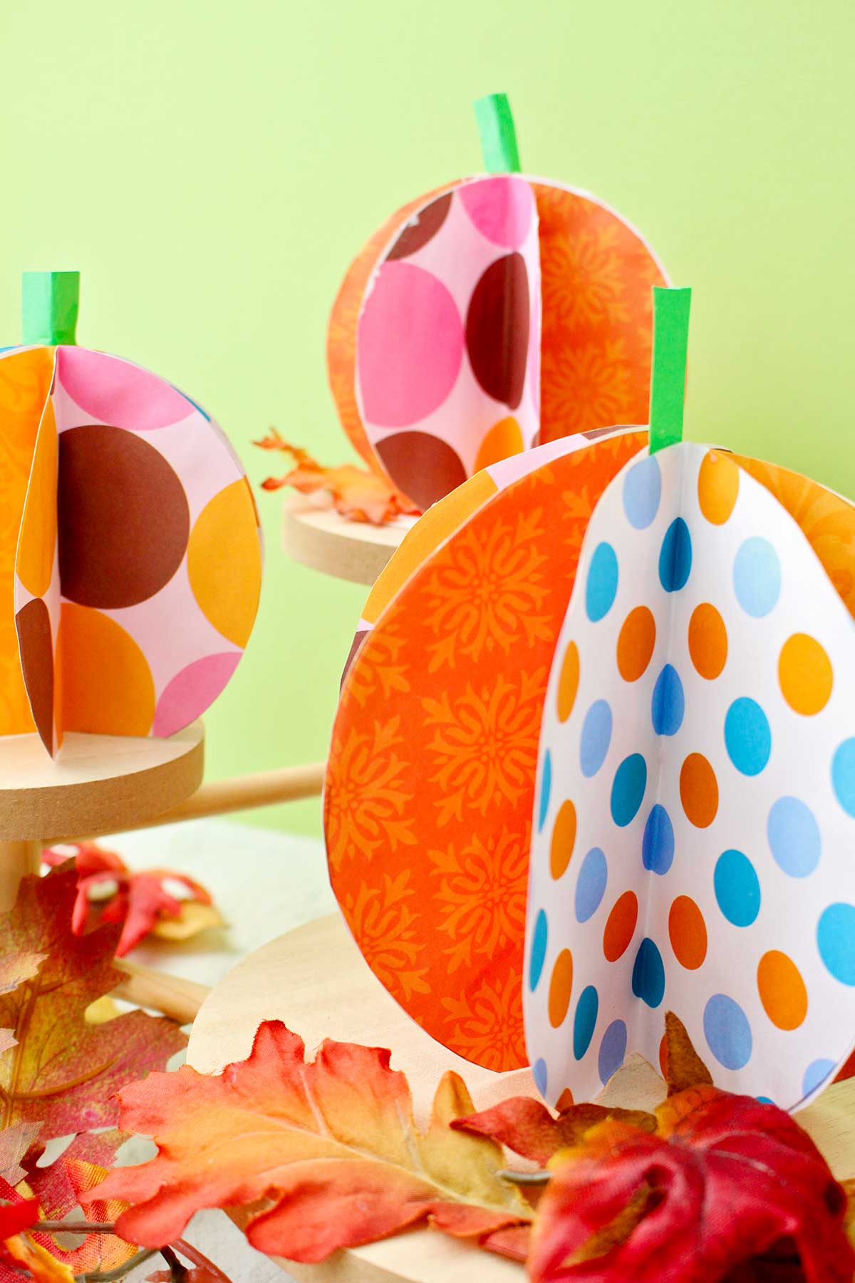 Three colorful paper pumpkins on wooden pedestals with fake leaves decorating near by.