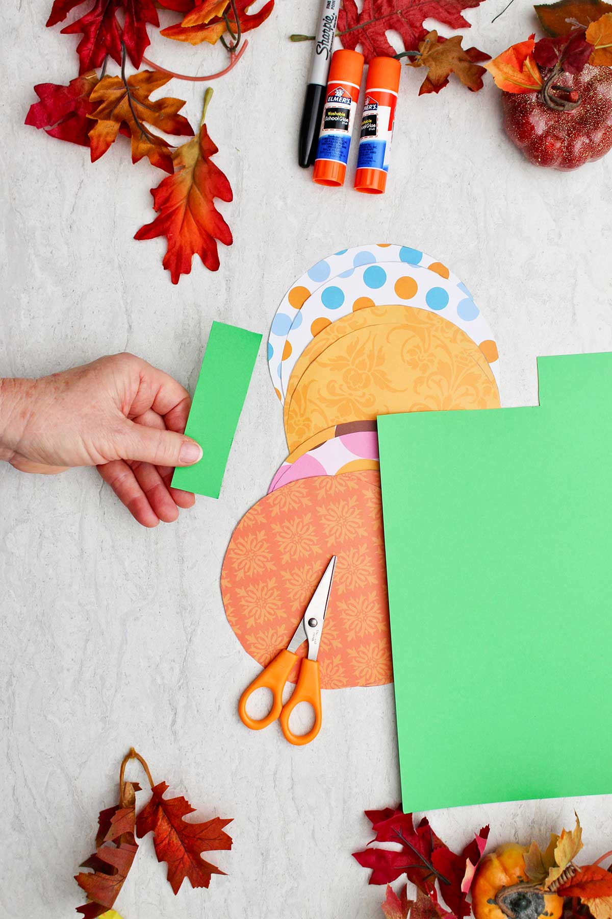 A person holding a green rectangle for a stem for the paper pumpkin with circles of colorful patterned paper and decorations near by.