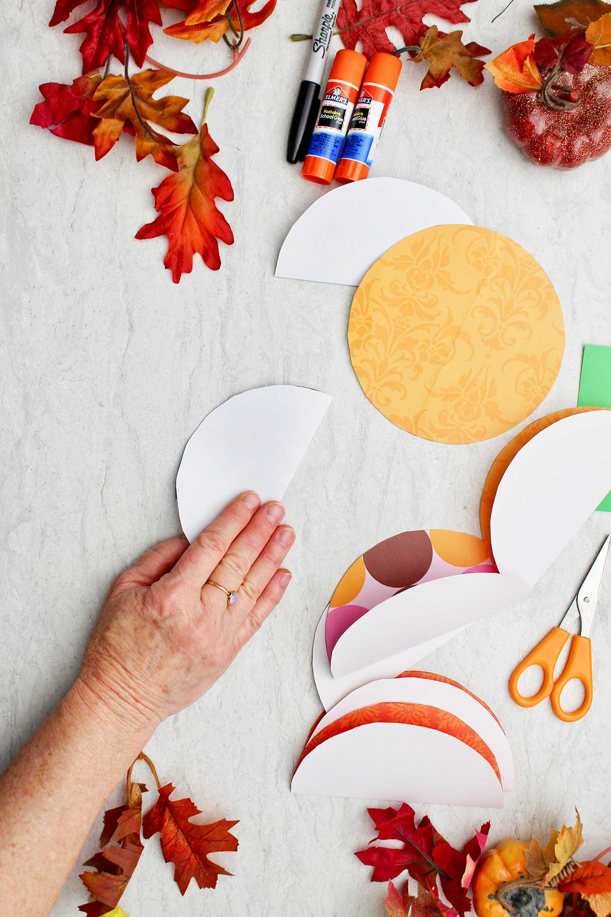 A person folding paper circles in half with fall leaves and supplies near by.