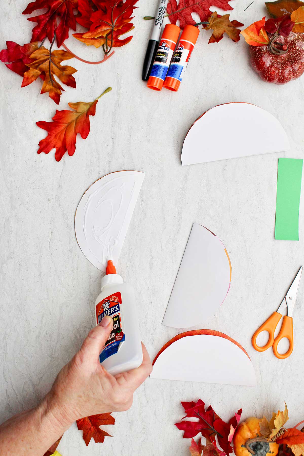 A person putting glue on a cut half circle of paper to make paper pumpkin with fall leaves and supplies scattered around.