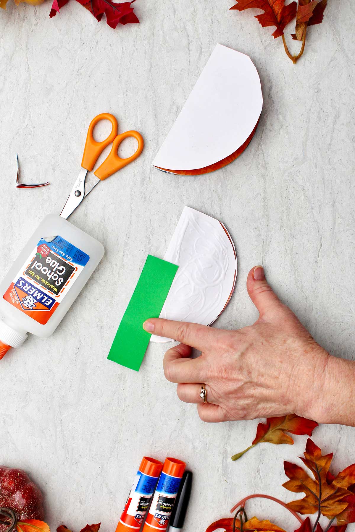 A person pasting a green construction paper stem on a folded half circle with supplies and leaves near by.