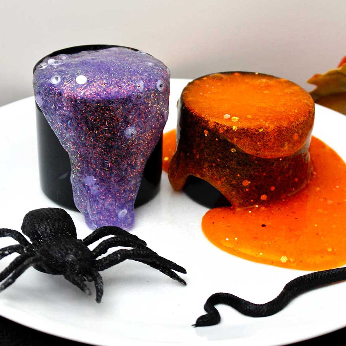 Two different spooky glittery slime recipes spilling out of their black containers with a fake spider near by.
